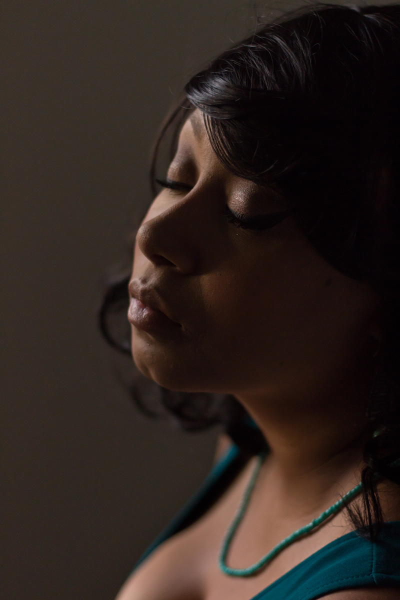 A black woman in her 20s is shown in this natural light portrait. She has medium length black hair and is wearing a dark teal blue dress with deep v-neck, brown leather belt, and blue beaded necklace. She identifies as plus size.
