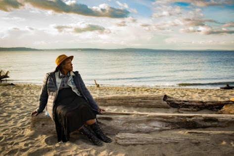 A plus-size woman of African-American ethnicity relaxes on a driftwood log on a sunny evening. She's wearing a brown hat, puffy black jacket, denim jean jacket, earrings, and black top and skirt. Behind her are a sandy beach, small waves, water and low mountains in the distance.