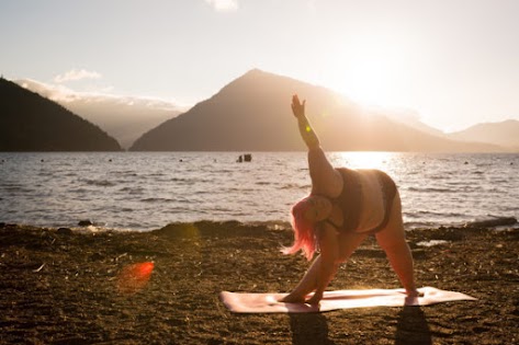 A Caucasian woman in her 30s who wears plus size clothing does yoga poses on a lake shore in bright, golden evening light. She has bright pink hair and pale skin, and is wearing workout gear that consists of a burgundy cropped top and briefs. Her yoga mat is a bright warm pink. She's very flexible and strong, and is an excellent example of confidence and positive body image. Lighting: natural light, high key, high contrast, sunset Environment: lake, shore, outdoors, mountains, nature, wilderness, water Mood: confident, happy, strong Themes: flexibility, strength, yoga, fitness, exercise, health at every size, joyful movement, positive body image, body confidence, body positive Ethnicities, genders and abilities of note: Caucasian Location: Olympic Mountains, Washington State, Pacific Northwest, United States